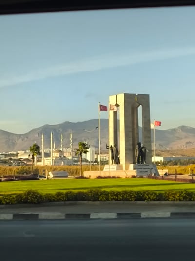 The image depicts a monument situated within a landscaped roundabout in Gonyeli, Northern Cyprus. The central feature is a large, light-colored monument composed of three vertical rectangular sections, with a central gap, resting on a multi-tiered base. Dark-colored statues of multiple human figures are positioned on either side of the monument's central structure. Two flagpoles are visible; one on the left prominently displays the flag of Turkey, while another on the right flies the flag of the Turkish Republic of Northern Cyprus. Inscriptions are present on the monument's base.

The roundabout itself features neatly trimmed green grass and low-lying green bushes. Two palm trees are visible within the roundabout, one to the left and another to the right of the monument. A streetlamp pole stands to the left of the monument.

In the mid-ground, beyond the roundabout, a large mosque with a central dome and four prominent minarets is visible. Various other buildings and urban structures can be seen interspersed among vegetation. The background features distinct mountains under a clear, light blue sky with minimal cloud cover.

The foreground shows an asphalt road surface with a painted curb featuring black and white stripes. The image is captured from a slightly elevated perspective, possibly from inside a vehicle, indicated by a dark horizontal bar across the top of the frame. The scene is illuminated by natural daylight, casting subtle shadows.