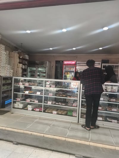 The image depicts the interior of a bakery in Krian, Indonesia. In the foreground, a young man wearing a red and black plaid shirt and black pants is standing at a display case filled with various pastries. He is facing a female employee wearing a black hijab and a grey top, who is standing behind the counter. To the left of the young man, another female employee wearing a red top and a headscarf is visible. Behind the counter, there are several refrigerators, one of which is a Coca-Cola branded cooler. The bakery is stocked with numerous boxes stacked high on shelves to the left of the scene. The display cases contain rows of individually packaged donuts and other baked goods, each with a price tag. The floor is tiled, and a ramp leads down from the entrance. The lighting is provided by overhead recessed lights.