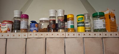 This is a close-up, eye-level shot of a collection of spice jars and food packaging displayed on a shelf. The items are arranged horizontally and are upside down, suggesting the photo was taken from below, perhaps looking up at a shelf attached to a wall. The background consists of a tiled wall with a decorative border.
The objects on the shelf are predominantly spices and baking ingredients. Visible items include a yellow Knorr "Aromat" seasoning blend, a Santa Maria spice mix for chicken, steak, onion, paprika, and garlic, and a bottle of "SPICE MIX WITH GARLIC". There are also containers of what appear to be dried herbs, chili flakes, and baking powder ("BAKPULVER LEIVINJAUHE" from Dr. Oetker). A blue package of "VANILLINSOCKER" (vanilla sugar) from Belbake is also present. To the far left, a portion of an orange cardboard box labeled "SUNNUNTAI" is visible.
The overall scene is that of a well-stocked spice rack or pantry shelf. The lighting appears to be artificial and even. There are no people or discernible weather conditions. The text on the products indicates a European context, with Finnish and German visible, and the location context provided is Orimattila, Finland. The image focuses on the product labels and their arrangement.