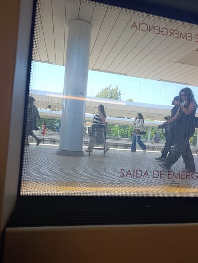 The image shows a train station platform in Pinhal Novo, Portugal, viewed through a window. People are walking on the checkered platform. A large white pillar stands prominently on the platform. In the background, trees and a blue sky are visible. The words "SAIDA DE EMERGENCIA" (Emergency Exit in Portuguese) are written in red on the platform. Another sign in the distance reads "PINHAL NOVO". The lighting suggests it is daytime with bright sunlight.