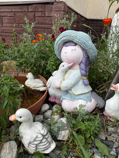 In the city of Zmeinyaya Gorka Ks, Russia, a garden scene is depicted. The foreground features several decorative ceramic figures of ducks and a figurine of a girl with purple hair and a blue knitted hat, holding a white duck. The girl is seated, her body in a light blue jacket with gold trim, and a light pink lower body. The ducks are positioned around the girl, one in a brown pot filled with water, and two on the ground. The background consists of a red brick wall and various plants, including greenery, small red and orange flowers, and some dry stalks. The ground is covered with dark gray gravel and scattered rocks. The overall setting suggests an outdoor garden space.