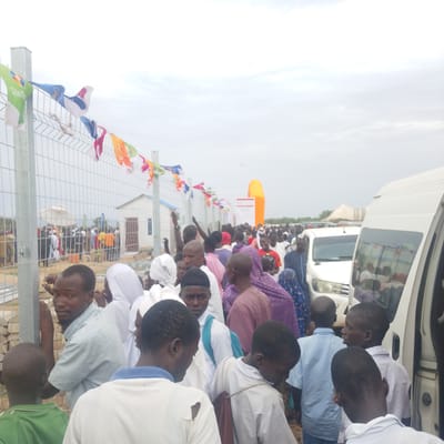 This is a photograph of a gathering of people in Wuro Ngangandi, Nigeria. The scene appears to be outdoors, with a cloudy sky overhead. In the foreground, a man with a beard and a light blue shirt is leaning against a metal fence. Behind him, several other individuals are visible, including a person wearing a white hijab and another wearing a white shirt. The fence itself is adorned with a string of colorful triangular flags, some of which have logos or text.

Beyond the fence, a crowd of people is milling about. A small white building with a blue roof is situated in the middle ground, and a large orange inflatable structure is visible to the right. Several vehicles, including white vans, are present in the background, suggesting transportation for the attendees. The overall impression is that of a public event or gathering.