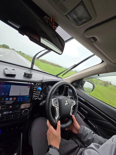 An interior shot of a car shows a driver's perspective on a rainy day in what appears to be Бабстово, Russia. A person, wearing a grey jacket and dark pants, holds the Mitsubishi-branded steering wheel, suggesting they are driving or about to drive. Raindrops are visible on the windshield and side mirror, with the wipers in an upright position, indicating wet weather conditions.
Through the windshield, a green field borders a wet asphalt road, which stretches into the distance under an overcast sky. Inside the vehicle, a large touchscreen infotainment system on the dashboard displays "12:41," a navigation map, and "87.5 FM" radio with the song title "Чужая свадьба" (Someone else's wedding) and "Я. Музыка" (Yandex.Music). The climate control shows "25.0" degrees Celsius, "DUAL," and "ECO MODE." Other visible text includes "MITSUBISHI" and "SRS AIRBAG" on the steering wheel, and "DIOGEスーパー" (DIOGE Super) on a car air freshener/diffuser on the dash. A small blue clip is attached to an air vent, and a dashcam or similar device is visible near the rearview mirror. The overall scene suggests a journey in rural Russia during daytime rain.