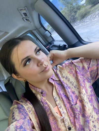 The image shows a woman sitting in a car, looking upwards and to the side. She has long dark hair pulled back and is wearing a light-colored shirt with a purple and yellow snake-like pattern. Her left arm is resting on her lap, and her right hand is near her face, touching her chin. The interior of the car is visible, including part of the ceiling and a window showing a blurred landscape of trees and water. Based on the provided context, the location is the city of Irganay, Russia.