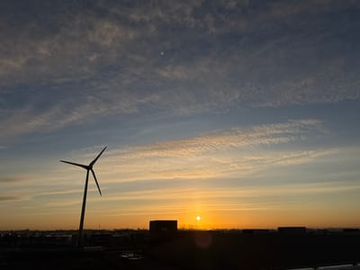 The image depicts an outdoor scene at either sunrise or sunset over the city of Naaldwijk, Netherlands. The sky occupies approximately two-thirds of the frame, transitioning from dark blue-grey in the upper portion with thin, wispy clouds, to light blue, yellow, and deep orange towards the horizon. The sun, appearing as a distinct orange circle, is positioned low on the horizon, slightly right of the frame's horizontal center. A smaller, brighter circular artifact is visible directly above the sun, and a faint, greenish circular artifact is present in the upper-left quadrant of the sky. Multiple thin, white linear contrails are present in the middle and upper-right sky.
The lower third of the image depicts a dark, silhouetted urban or industrial landscape. A prominent three-bladed wind turbine is silhouetted on the left, its dark form contrasting against the bright sky. Below the horizon, numerous dark, block-like structures, indicative of buildings, form an urban or industrial skyline. One distinct rectangular building is positioned to the left of the sun. A horizontal line, possibly a road, is discernible in the mid-ground, with a light-colored vehicle (possibly a truck) positioned to the right of the wind turbine's base. The immediate foreground shows dark, irregular shapes consistent with wet ground or puddles. No individuals are visible.
