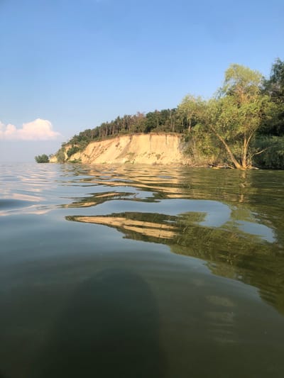 The image depicts a serene natural scene featuring a body of water in the foreground and a cliffside covered with trees in the background. The water is calm, with reflections of the cliff and sky visible on its surface. The cliff is characterized by its light-colored, sandy soil and is topped with a dense forest. The sky above is a clear blue, with a few wispy clouds. The scene appears to be a peaceful, natural landscape, likely a rural area or a park. The specific location in Nahirne, Ukraine, is a coastal area or lakeside, given the presence of water and the cliff formation. There are no people or discernible actions taking place in the image.