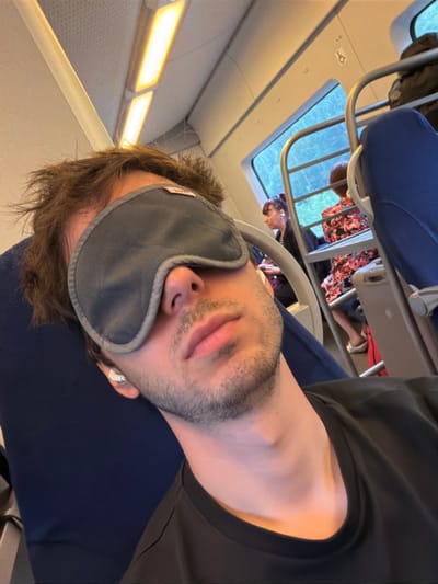 A man wearing a dark gray eye mask and white earbuds is asleep on a train. He is seated next to a blue upholstered seat and is wearing a black t-shirt. The train car's interior is visible, with overhead lighting and a window showing blurred greenery outside. Other passengers are partially visible in the background, one in a floral pattern top and another with dark hair. The scene appears to be on a typical commuter or intercity train. There is no specific indication of the city of Drezna, Russia, in the image.