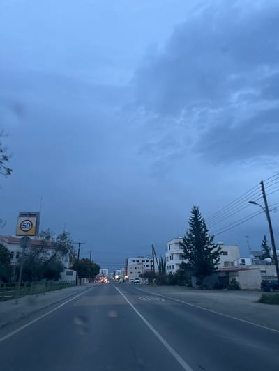 The image presents a street view in Latsia, Cyprus, under a dark blue-grey, uniformly overcast sky. The perspective is from a vehicle on a straight asphalt road, which displays a dashed white center line, a solid white line on the right shoulder, and a pedestrian zebra crossing in the immediate foreground. A circular yellow speed limit sign, marked "50," is visible on the left side of the road. Buildings of multi-story construction, predominantly light-colored, line both sides of the thoroughfare. On the left, a prominent building supports a large sign structure. On the right, a tall, slender coniferous tree stands near a multi-story building, with overhead power lines extending along utility poles. Distant streetlights are illuminated. Multiple vehicles are visible in the distance, either moving or parked, some displaying active lights. A single individual is observed walking on the right side of the road, moving away from the camera. The road surface shows reflective patches, indicating dampness.