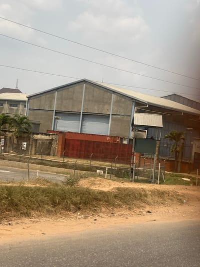 This image depicts an industrial scene in Odo Ona, Nigeria. In the foreground, an asphalt road runs horizontally, bordered by a strip of dry earth and sparse, yellowish-brown grass. Beyond this, a chain-link fence topped with barbed wire delineates a property line.
Behind the fence, a prominent, large industrial building with a grey concrete facade and a light-colored corrugated metal roof dominates the midground. Two large, grey roll-up garage doors are visible on its front. Steel support beams are vertically integrated into the facade. To the left and slightly behind, another industrial building with a similar roofline and small rectangular windows is partially visible. An attached structure with a corrugated metal roof extends from the right side of the main warehouse.
In front of the main warehouse and behind the fence, a large, weathered, reddish-brown shipping container is positioned horizontally, with faint markings "MSC 401" visible on its side. To the right of the container, a dark green rectangular panel is mounted on a grey metal frame structure. Palm trees are visible within the fenced property. Overhead, multiple thin power lines traverse the sky, which is a pale, overcast grey. No individuals or active interactions are discernible.