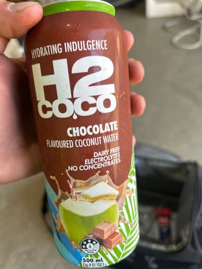 A person holds a brown bottle of H2coco Chocolate Flavoured Coconut Water. The label states it's a "hydrating indulgence," "dairy-free," and contains "electrolytes" and "no concentrates." The can's design includes an image of a coconut with chocolate pieces and palm leaves. A four-star health rating is visible, along with the volume, which is 500 ml (16.9 fl oz). A black gym bag sits in the background.
