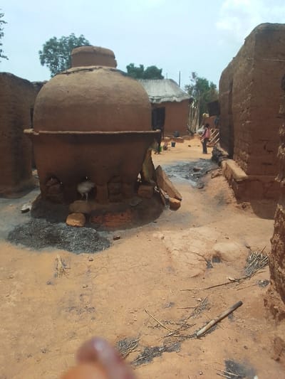 The image shows a large, round, clay oven in the center of the scene. The oven has a wide base and is supported by brickwork. A white chicken stands on a rock in front of the oven. The oven is located in a narrow, dirt alleyway between mud-brick buildings. The alleyway is dusty and littered with twigs and other debris. There is a small amount of black ash on the ground in front of the oven. In the background, two people can be seen walking in the alley. They appear to be on their way to or from the oven. The buildings in the background are made of mud brick and have thatched roofs. The sky is clear and blue, and the sun is shining. The scene is located in the village of Kwal, Nigeria, at the coordinates 9.804 latitude and 8.647 longitude.