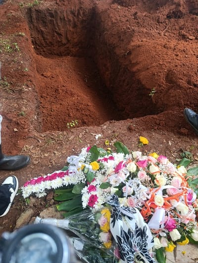 The image depicts an open grave site in Muthetheni ward, Kenya. In the foreground, a substantial collection of funeral floral arrangements is visible on disturbed reddish-brown earth. These include a cross-shaped wreath composed of white chrysanthemums and red roses, alongside a large, mixed arrangement featuring various roses in shades of pink, white, yellow, and peach, accented with white baby's breath and multicolored ribbons. Several bouquets in clear plastic wrap are also present. The ground itself is reddish-brown soil, with sparse green vegetation. Partially visible human feet are present around the floral tributes and grave: a left foot in a black and white sneaker, a left foot in a black rubber boot (gumboot), and a right foot in a black dress shoe. In the midground, a rectangular grave is dug into the reddish-brown earth, approximately 2 meters long with steep, unreinforced walls, appearing to be at least 1.5-2 meters deep. The bottom-center of the frame shows the blurred top of a camera lens. The scene suggests an active burial preparation or ceremony.