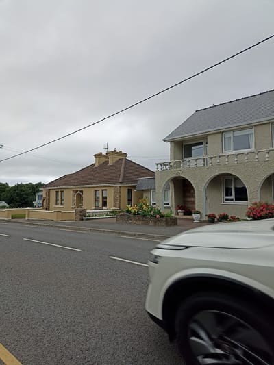 A front-left view captures a white vehicle driving on a paved road in Annagry, Ireland. The road features white lane markings and a yellow line on the left edge. Adjacent to the road are two houses. The house on the left is a single-story structure with a brown, tiled roof, light yellow walls, and stone accents. It has several windows and a low, beige-colored wall in front. The house on the right is a two-story building with a grey, tiled roof, a stucco exterior, and arched openings on the ground floor. A balcony with a white railing runs across the second story. Both houses have flower beds with red and yellow blooms. The sky is overcast and grey, and a power line runs diagonally across the upper portion of the image. Trees are visible in the background.
