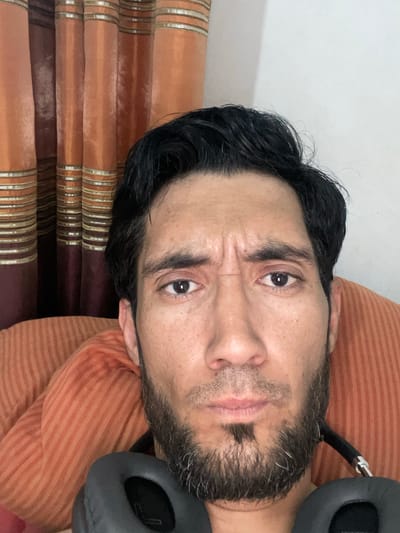 The image is a close-up, front-facing selfie of a man with dark hair and a beard. He is looking directly at the camera with a slightly furrowed brow and his mouth closed. He is wearing dark headphones around his neck. The background consists of orange and brown striped curtains. The image does not provide any information about the city of Mashrab, Uzbekistan.