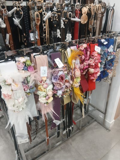 The image displays a retail display of various belts, primarily decorative elastic belts adorned with artificial flowers and feathers, as well as some plain leather and braided belts. The belts are organized on multiple tiered metal racks. The foreground shows a close-up of the decorative belts, highlighting their floral and feathery embellishments in a variety of colors, including pink, white, blue, red, and yellow. The background reveals more belts, some with elaborate buckles. The scene appears to be an indoor retail environment, likely a clothing or accessories store. The flooring is light gray tile. There are no people visible in the image. The image does not provide any specific information or context that would indicate its location in the city of Ávila, Spain.