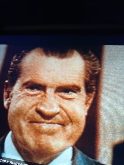 The image is a close-up, slightly grainy photograph of a man's face, appearing to be a still from a video or television broadcast. The man has fair skin, thinning brown hair, and is smiling broadly, showing his teeth. He is wearing a white collared shirt and a dark suit jacket. The background is out of focus and appears to be indoors with some subtle lighting variations. The bottom left corner of the image contains text in a foreign language, possibly Russian, that is partially visible. There is no discernible connection to Mountcharles, Ireland.