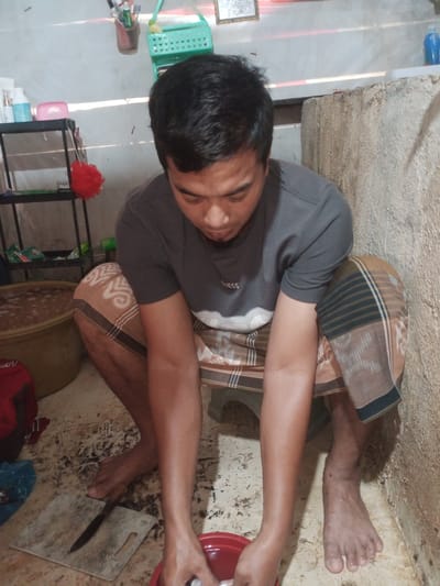 In a dimly lit, possibly outdoor or semi-enclosed area in Cotseutui, Indonesia, a man is crouched down, appearing to be preparing food or cleaning something. He is wearing a dark gray t-shirt with a white "GUESS" logo and a traditional sarong with a brown, beige, and black checkered pattern. His focus is directed downwards at his hands, which are submerged in a red plastic basin. To his left, a large, light brown tub filled with a liquid substance is visible. In front of him, a worn wooden cutting board with a knife resting on it lies on the concrete floor. The floor is stained and appears wet in places. To the left of the man, a black shelving unit holds various items, including bottles and a red loofah-like object hanging from it. The background consists of a rough, textured concrete wall and a lighter-colored wall with some shelving and decorative items.