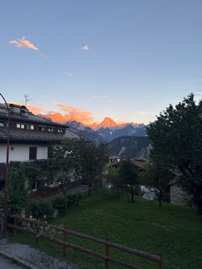 This is a photograph taken at sunset in the vicinity of Valle di Cadore, Italy. The image captures a mountainous landscape illuminated by the warm, orange hues of the setting sun, with snow-capped peaks prominently featured. In the foreground, a residential building with a dark roof and white walls is visible, featuring balconies and windows. Trees and a grassy lawn are present in front of the building, with a rustic wooden fence bordering a paved area. The sky is clear with a few scattered clouds tinted pink by the sunset. The overall scene depicts a tranquil mountain village during twilight.