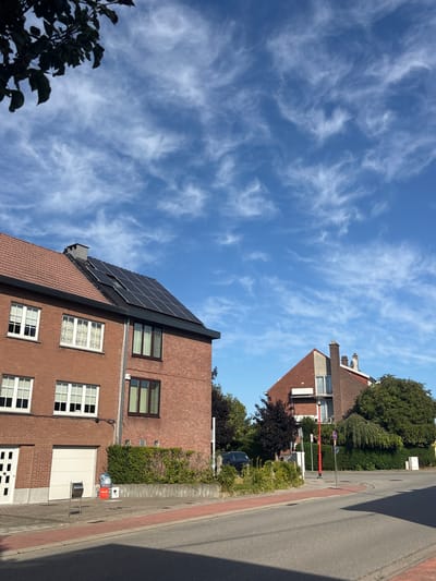 The image depicts a residential street scene in Strombeek-Bever, Belgium, on a clear day with wispy white clouds in a blue sky. In the foreground, a paved road with a red border runs diagonally from the bottom right towards the center. To the left of the road is a sidewalk.
Dominating the left side of the image is a two-story brick house. The left section of the house has a terracotta tiled roof and features several white-framed windows. A white garage door is visible on the ground floor. Adjacent to this, the roofline shifts, and solar panels are installed on a darker, sloped section of the roof. This section of the house has a chimney. To the right of this, another brick house with similar windows is visible.
Behind the houses, lush green trees and bushes form a dense barrier. A dark-colored car is parked on the left side, partially obscured by foliage. Several street signs and a red lamppost are positioned along the sidewalk near the corner of the street. The overall impression is a quiet suburban street.