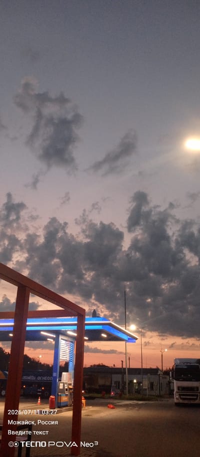 A gas station is pictured at dusk in Mozhaysk, Russia. A white truck is parked to the right of the frame, and a gas pump is visible under a brightly lit canopy. The sky is filled with dramatic clouds, and the sunset colors are visible on the horizon. The timestamp "2026/07/11 03:27" and "Можайск, Россия" are overlaid on the image, along with the text "Введите текст" and the TECNO POVA Neo³ logo.