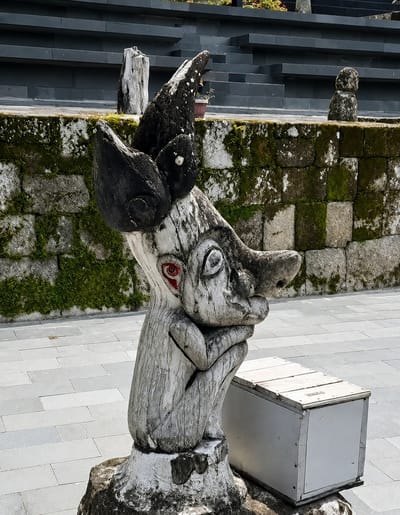 A weathered wooden sculpture of a creature with a long snout and red eyes sits on a stone base in Ambarita, Indonesia. The sculpture is positioned next to a simple wooden bench. Behind the sculpture, a moss-covered stone wall is visible, with a dark, multi-tiered structure in the background. The ground is paved with light-colored rectangular stones.