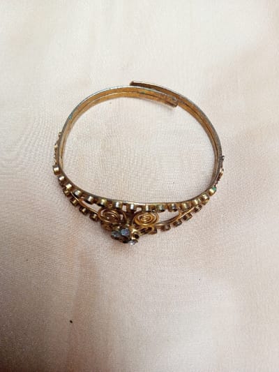 A gold-toned metal bracelet, appearing aged with visible green oxidation (verdigris) on its surface and adjustable clasp, rests on a light beige textile surface. The bracelet is an open-ended bangle design, featuring small, discolored stones embedded along its lower edge. A central ornamental section includes two filigree scrolls bordering a cluster of faceted stones, comprising a larger blue-green stone and several smaller clear or tarnished stones. The textile surface exhibits subtle texture and minor folds. No other objects or individuals are present. The scene is located in Qarabucaq qəsəbə, Azerbaijan.
