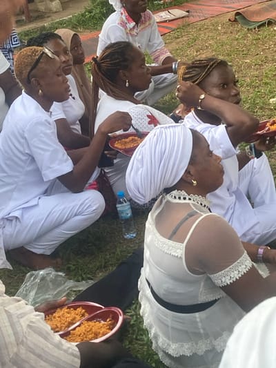 A group of African women, mostly in white, are seated outdoors. Many are eating from red plastic plates. The attire ranges from traditional white wraps to modern white lace tops. Some women have braided hair, others wear headscarves. A water bottle is visible amongst them. The scene appears to be a casual gathering, possibly a celebration or religious event.
