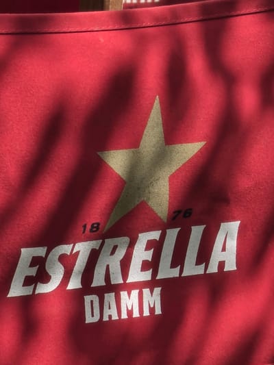 A close-up shot shows a red fabric, likely a shirt or apron, with a large, golden star emblem centrally placed. The words "ESTRELLA DAMM" are printed in white lettering below the star. To the left of the star, the number "18" is visible in black, and to the right, the number "76" is also in black. The fabric is textured and appears to be illuminated by natural light, casting shadows from what appears to be foliage across the surface. The scene is set in Magaluf, Spain.