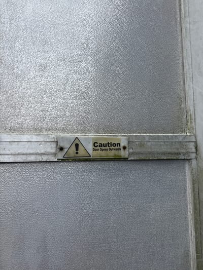 A close-up shot shows a section of what appears to be a door or wall with a textured, slightly reflective gray surface. A vertical metal strip runs down the center, where a caution sign is affixed. The sign, oriented vertically, reads "Caution Door Opens Outwards" above a triangular warning symbol containing an exclamation point. There are visible signs of wear and grime on both the metal strip and the sign. The image does not contain enough information to determine the specific location within Igwuruta, Nigeria.