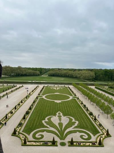 The image shows an aerial view of the formal gardens of the Chateau de Chambord, located in the Loire Valley of France at latitude 47.616, longitude 1.518. A large, manicured lawn with a patterned design extends from the foreground towards a line of trees. The design is outlined with gravel pathways, and there are rows of trees lining the pathways on either side. The background is a wide open field with trees on either side, which appear to be along the banks of a river, before the distant, forested horizon. A few people are walking on the gravel paths, which are lined with small trees and bushes. The sky is overcast with gray clouds. The photograph appears to have been taken from a window on one of the upper levels of the chateau, possibly the roof.