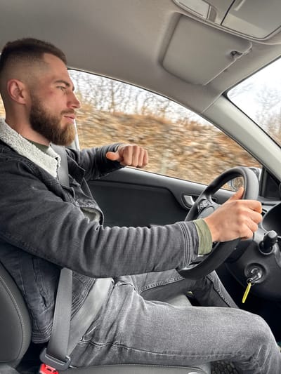 A male individual is observed in the driver's seat of a passenger vehicle, appearing to be in motion. He has short dark hair with a fade and a full beard, and is wearing a dark grey denim jacket with a light-colored sherpa lining, an olive green long-sleeved shirt, and dark grey jeans. A seatbelt is correctly fastened across his chest and lap. His left hand is positioned on the black steering wheel, while his right arm is bent, and his right hand forms a loose fist, resting near the open window or door panel. The vehicle's interior features a dark dashboard, a light grey headliner, and a sun visor in the upright position. Keys with a yellow fob are visible in the ignition. A faint reddish mark is present on his right cheekbone. The exterior environment, blurred due to motion and visible through the windows, consists of a natural landscape with bare trees and brownish ground cover, consistent with a non-foliage season. The specific location within Barutana, Montenegro, cannot be definitively identified from the image, but the setting is consistent with a road in a rural or semi-rural area.