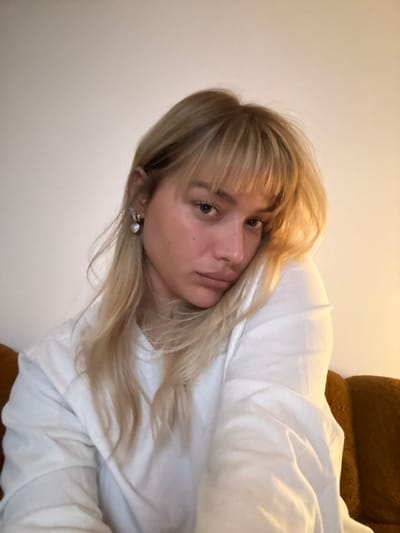 A woman with blonde hair and bangs is looking directly at the camera. She is wearing a white sweatshirt and appears to be lying down, possibly in bed, as a white blanket is draped over her. A brown cushion is visible in the background. She has an earring in her left ear. The image has been rotated, causing the perspective to be slightly tilted.