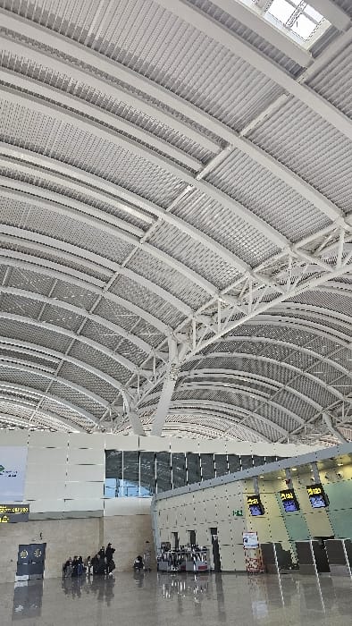 The image depicts the interior of a modern airport terminal, specifically at Houari Boumediene Airport in Dar El Beïda, Algeria. The scene is dominated by a high, arched ceiling constructed from white corrugated and perforated metal panels supported by exposed white metal trusses and beams. Integrated skylights are visible in the upper sections of the ceiling.
The floor is a polished, light-toned surface exhibiting strong reflectivity, mirroring the overhead structure and ambient light. The walls consist of large, light-colored modular panels and sections of dark-tinted glass windows, some reflecting the exterior sky.
In the mid-ground, a group of approximately 5-7 individuals is gathered on the left, mostly seated on the floor or low structures, surrounded by various pieces of luggage. They appear to be waiting. To the right, a series of service or check-in counters are visible, featuring angled digital display screens showing yellow and white alphanumeric information against a dark background. Metallic structures, likely baggage processing equipment, are positioned in front of these counters. A single figure stands near the counters on the right. A large white wall section with a green graphic is partially visible on the far left, above an unreadable dark electronic display. A dark doorway is present near the left edge of the frame. The overall environment is brightly lit.