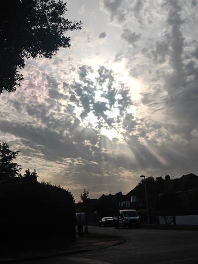 The image depicts a street scene in Worthing, United Kingdom, under a cloudy sky with the sun partially visible and casting crepuscular rays. In the foreground, a dark, silhouetted hedge occupies the left side of the frame. A paved road curves from the bottom left towards the center. On the right side of the road, houses with tiled roofs are visible in the midground, partially obscured by trees. A lamppost stands beside the road. Two vehicles are parked on the road: a black car on the left and a white van on the right. A person is walking on the sidewalk to the right of the white van, and another person is seen walking on the far right sidewalk. The sky is filled with scattered clouds, with a bright area where the sun is breaking through.