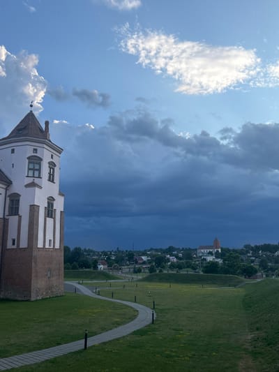 The image shows a view of a white and red brick tower on the left, part of a historical complex, likely a castle, situated in Жухавіцкі сельскі Савет, Belarus. The tower has a red, pitched roof with a small spire. In the background, a town or village is visible, characterized by scattered buildings with red roofs and green trees. A prominent red-roofed church with a tall tower stands out among the buildings in the distance. The foreground consists of a well-maintained lawn with a winding paved pathway and several lampposts. The sky is a dramatic mix of bright blue with fluffy white clouds in the upper portion, transitioning to dark, stormy clouds in the lower half, suggesting an approaching storm or the aftermath of one. The overall scene depicts a historical architectural site within a rural Belarusian setting under dynamic weather conditions.