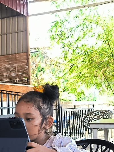 The image shows a young girl with her hair in a bun, secured with a yellow scrunchie, holding a dark-colored tablet. She is looking at the screen. She is wearing a striped shirt. The background shows a metal roof structure and lush green foliage with a wooden fence. A table and chair with an intricate back design are partially visible. The location is likely Matang, Indonesia, based on the prompt's information.