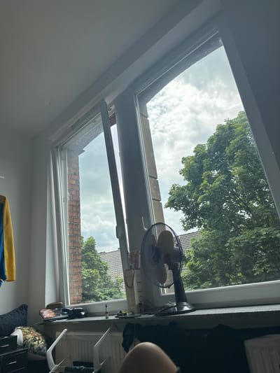 The image is a photograph taken from inside a room looking upwards towards a partially open skylight. The scene depicts an interior space with white walls and a ceiling. Through the skylight, a cloudy sky is visible, with large green trees and the rooftops of houses outside. A black oscillating fan is positioned on the windowsill of the skylight, angled towards the interior. Several small objects are placed on the windowsill and a shelf below, including what appears to be a book with visible text, eyeglasses, and a tablet device. The angle of the shot suggests the photograph was taken from a low vantage point, possibly from a bed or couch. The lighting indicates it is daytime, with diffused light from the overcast sky. The overall impression is a cozy, lived-in room with a connection to the outdoors through the open skylight. The location context is Hamm, Germany.