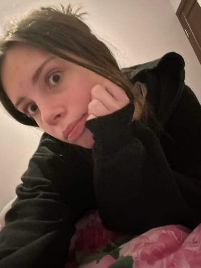 A selfie shows a person with fair skin, brown hair, and brown eyes. The person is wearing a black hooded sweatshirt. Their face is tilted, and their left hand is touching their cheek. A hint of pink floral bedding is visible in the upper right corner. The background is a plain, light-colored wall, with a hint of a wooden cabinet visible at the top.