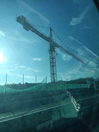 A construction crane stands against a bright blue sky with wispy clouds. The crane is yellow and orange, with a long jib extending horizontally. Below the crane, a construction site is visible, covered with green netting. Trees line the horizon behind the netting. The image appears to be taken through a window, as reflections of an interior are visible on the glass. There are no people or animals clearly visible in the image. The location is specified as Hrusice, Czechia.