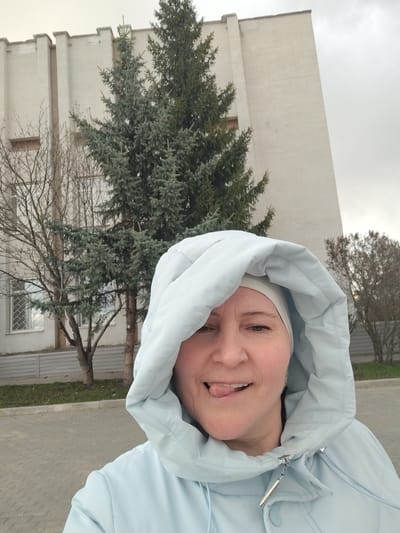 A woman is captured in a close-up selfie outdoors, wearing a light blue hooded jacket with the hood pulled up over her head. She also has a white or light-colored head covering visible beneath the hood. Her expression is playful, featuring a wink with one eye and her tongue slightly sticking out.
The background shows a large, light-colored building with vertical architectural elements or columns. To the left, a bare deciduous tree stands in front of the building, while a tall, dense evergreen coniferous tree (resembling a spruce or fir) is more centrally located behind the woman. On the right side, another smaller, bare shrub or tree is visible in the distance. The ground beneath is a paved area made of rectangular pavers, likely a sidewalk or pedestrian path. The sky is overcast and gray.
The image depicts a casual, lighthearted moment, suggesting the woman is enjoying her time outdoors, possibly on a cool or chilly day given her attire. The overall setting points to an urban or semi-urban environment in Yanaul, Russia. No text is visible in the image.