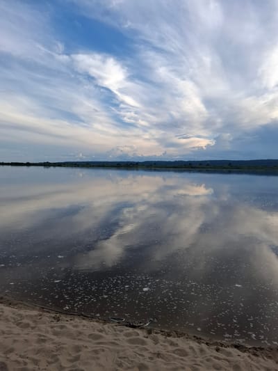The image shows a wide, calm river reflecting a dramatic sky filled with wispy clouds. The foreground features a sandy bank with some debris, including a branch and what appears to be a small piece of driftwood or a shell. The distant shore is lined with trees, creating a dark, silhouetted horizon. The lighting suggests it is late afternoon or early evening. This scene is located in Malaya Sazanka, Russia.