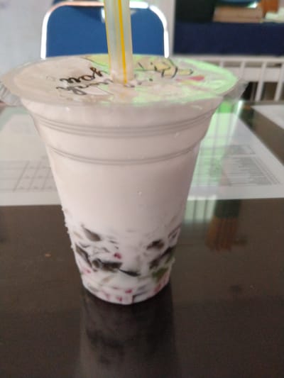 A tall, clear plastic cup filled with a milky beverage containing various colorful jellies and possibly fruit pieces sits on a dark, reflective surface. A yellow and white striped straw protrudes from the sealed lid of the cup. The lid has faint text and a colorful design. In the blurred background, a blue chair and a white railing are visible, suggesting an indoor setting, possibly a cafeteria or dining area in Pulorayeuk, Indonesia.