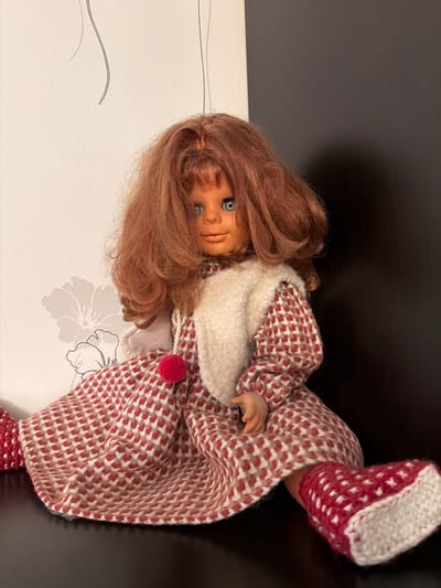 This is a close-up, rotated photograph of a doll. The doll has reddish-brown, wavy hair that falls around its face. It has blue eyes and a doll-like facial expression. The doll is wearing a red and white checkered outfit, which appears to be a jumpsuit, and has matching booties. Around its neck, the doll is wearing a fluffy white scarf. A small red pom-pom is attached to the outfit near the chest area. The doll is positioned on a surface that transitions from a white patterned background to a dark, reflective surface. The white background has a subtle floral design. The lighting suggests an indoor setting, likely a room. No specific time of day, weather, or emotional cues are evident beyond the doll's appearance. No visible text is present in the image. The location context of Maloyaroslavets, Russia, is provided but not visually discernible from the image itself.