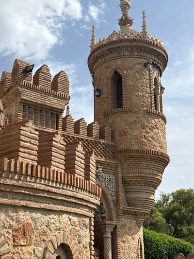 This is a low-angle, vertical shot of a whimsical, castle-like structure made of stone and brick. The building features multiple towers with decorative elements, including crenellations, pointed spires, and arched windows. The brickwork is intricate, with various patterns and textures, particularly on the curved sections of the towers. Small lights are attached to some of the brick structures. In the background, a bright blue sky with scattered clouds is visible, along with lush green trees and foliage peeking from the top right of the frame. The lighting suggests it is daytime, likely late morning or early afternoon, given the brightness and shadows. The overall impression is one of an unusual, fairytale-like architectural design in an outdoor setting. The location is Benalmádena, Spain. No text is visible in the image.