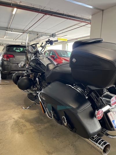 In a covered parking garage, a black Harley-Davidson motorcycle is parked facing away from the camera. The motorcycle has leather saddlebags and a large backrest. A grey car is parked behind and to the left of the motorcycle, and a red car is parked behind and to the right. The garage has a concrete floor and ceiling with exposed pipes and fluorescent lighting. A yellow sign with German text is visible on the ceiling. A license plate with "B" is visible on the back of the motorcycle. The image was taken in Weiswampach, Luxembourg.