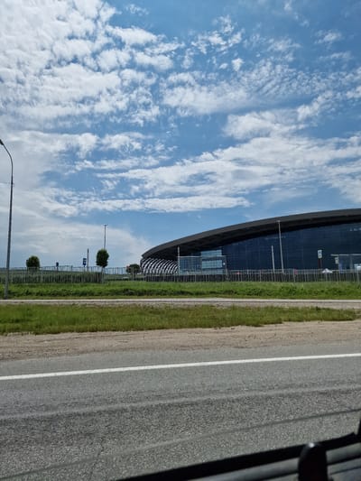 The image depicts a wide, outdoor shot of a modern airport terminal building in Krasny Pakhar, Russia. The terminal is characterized by its sweeping, dark roof and expansive glass facade, reflecting the partly cloudy blue sky above. In the foreground, a paved road with a white lane marking runs horizontally across the bottom of the frame, suggesting the perspective is from a vehicle. To the left of the road, a strip of green grass and a dirt embankment are visible. Beyond the grass is a metal fence, separating the roadside from the airport grounds. A few manicured trees are planted behind the fence. Several lampposts are also present, including a tall one on the far left and a shorter one near the center. The sky is filled with scattered white, wispy clouds that create a dynamic texture against the blue. The overall scene is bright and airy, indicative of daylight.