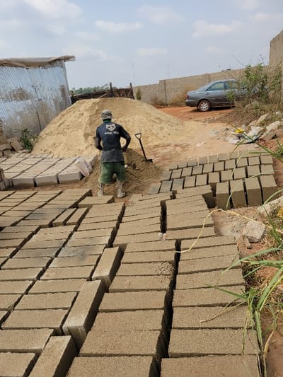 The image depicts an outdoor block-making operation in Nwofia, Nigeria, under a partly cloudy sky. In the foreground, numerous light grey-brown sandcrete blocks are arranged in parallel rows on reddish-brown earth, with some green vegetation visible on the far right. A male individual, seen from behind, occupies the center-midground. He wears a dark t-shirt with a white skull graphic on the back, dark shorts, and light-colored footwear, standing in front of a substantial mound of light-colored sand. A black shovel is partially inserted into a smaller sand pile near his feet. To the left of the sand mound, a structure composed of corrugated metal sheets is visible, alongside more rows of blocks and a white sack on the ground. In the mid-right background, a grey sedan is parked on an unpaved area, with a low wall constructed from concrete blocks extending behind it. The distant background consists of sparse vegetation under a hazy sky.