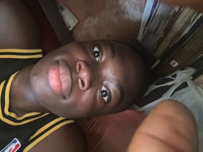 This image captures a close-up, angled portrait of a single individual in the city of Iriebe, Nigeria. The subject, a person with dark skin, is positioned as if lying down on their left side or back, looking directly into the camera. They are wearing a dark-colored garment with two parallel yellow horizontal stripes on the visible shoulder/upper arm area, and a partial NBA logo is discernible on the lower left of the garment. The individual possesses brown eyes and full, light pink lips. The immediate surface supporting the subject's head is a reddish-brown textured material. In the background, to the upper left, a white rectangular object, potentially an electronic device, rests on a light brown surface. To the upper right, a vertical stack of items, resembling books or woven panels, is visible; one item displays a "PAL" label on its edge. Below these stacked items, a crumpled light-colored material, consistent with fabric or plastic, is present. A blurry finger partially obscures the lower right foreground. The overall lighting of the scene appears subdued.