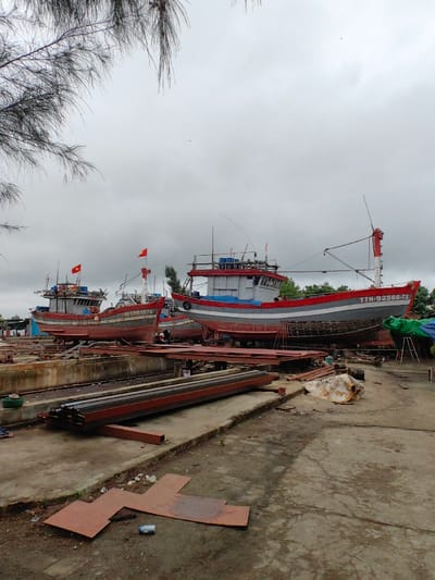 The image depicts a boat repair and construction yard in Thuận An Ward, Vietnam, under an overcast sky. Three large fishing vessels are the dominant subjects, elevated on dry-dock-like support structures.

The vessel in the right foreground is painted with a red top, a white stripe, and a gray hull, bearing the registration "TTH-92586-TS" on its bow. It features a gray cabin, a red mast, and various rigging. Another vessel, left-center, shares a similar color scheme with a red hull base and gray upper hull, marked "TTH-50612-TS". A third vessel is partially visible behind the second. All three boats prominently display the red Vietnamese flag with a yellow star. Blue tarpaulins are draped on and around the boats, indicating protection or ongoing work.

In the foreground, the ground consists of a rough concrete surface interspersed with dirt patches. Stacked horizontally are numerous dark red-brown metal beams or structural elements, positioned on or near a concrete track system. Smaller, irregularly shaped rusted metal plates lie flat on the ground. A discarded white plastic bottle is visible near these plates. Various debris and additional construction materials, including pipes and scrap metal, are scattered throughout the yard.

Overhanging dark, needle-like tree branches extend into the upper left of the frame. Green foliage and trees are visible in the background behind the vessels, bordering the industrial area. No individuals are visible in the scene, though the arrangement of materials and boats suggests active industrial operations.