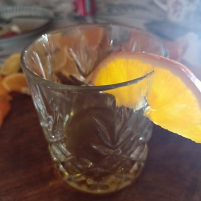 A close-up shot captures a glass of amber liquid, likely a cocktail or whiskey, garnished with a vibrant slice of orange. The glass itself appears to be made of cut glass, with a textured pattern that catches and refracts the light. The orange slice is placed artfully on the rim of the glass, its bright yellow-orange hue contrasting with the dark liquid inside.
In the background, out of focus, are hints of a domestic setting. The blurry elements suggest a table surface, possibly made of wood, and other objects that could be dishes or tableware. There are also glimpses of what appears to be a patterned fabric, perhaps a tablecloth or placemat. The overall lighting suggests a dimly lit environment, possibly indoors with natural light filtering in from a window, or an artificial light source. The bright highlights on the glass and the orange slice indicate a light source directly on them.
No people are visible in the frame. The focus is entirely on the drink and its presentation. The mood is calm and inviting, suggesting a moment of relaxation or enjoyment. There are no discernible signs or text within the image. The scene evokes a sense of simple pleasure, focusing on the aesthetic appeal of a well-prepared drink. The location context of Limerick, Ireland, does not provide any specific visual cues within the image itself, but it grounds the scene in a particular geographical and cultural setting.